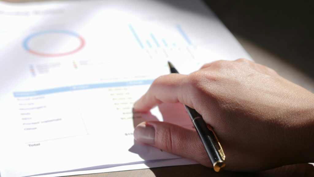 pexels-photo-7054416-7054416 A close-up view of a hand holding a pen and examining financial charts on paper.
